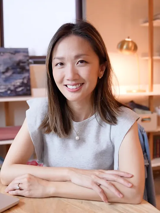 Smiling woman with shoulder-length brown hair wearing a sleeveless gray top and pearl necklace, sitting at a wooden table with arms crossed.