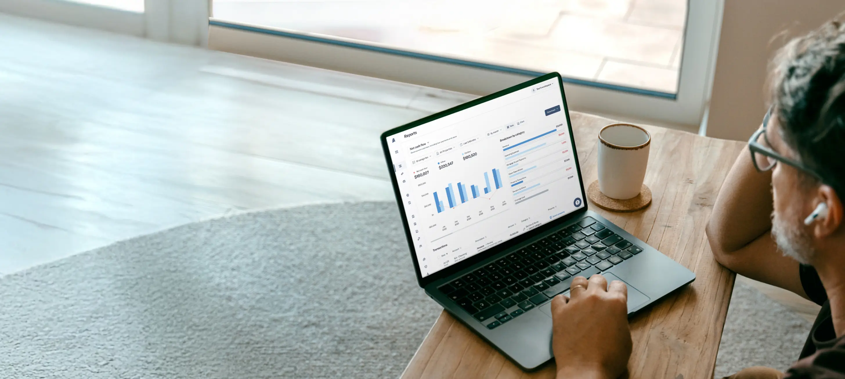 Person using a laptop on a wooden table displaying financial reports and charts, with a coffee cup nearby.