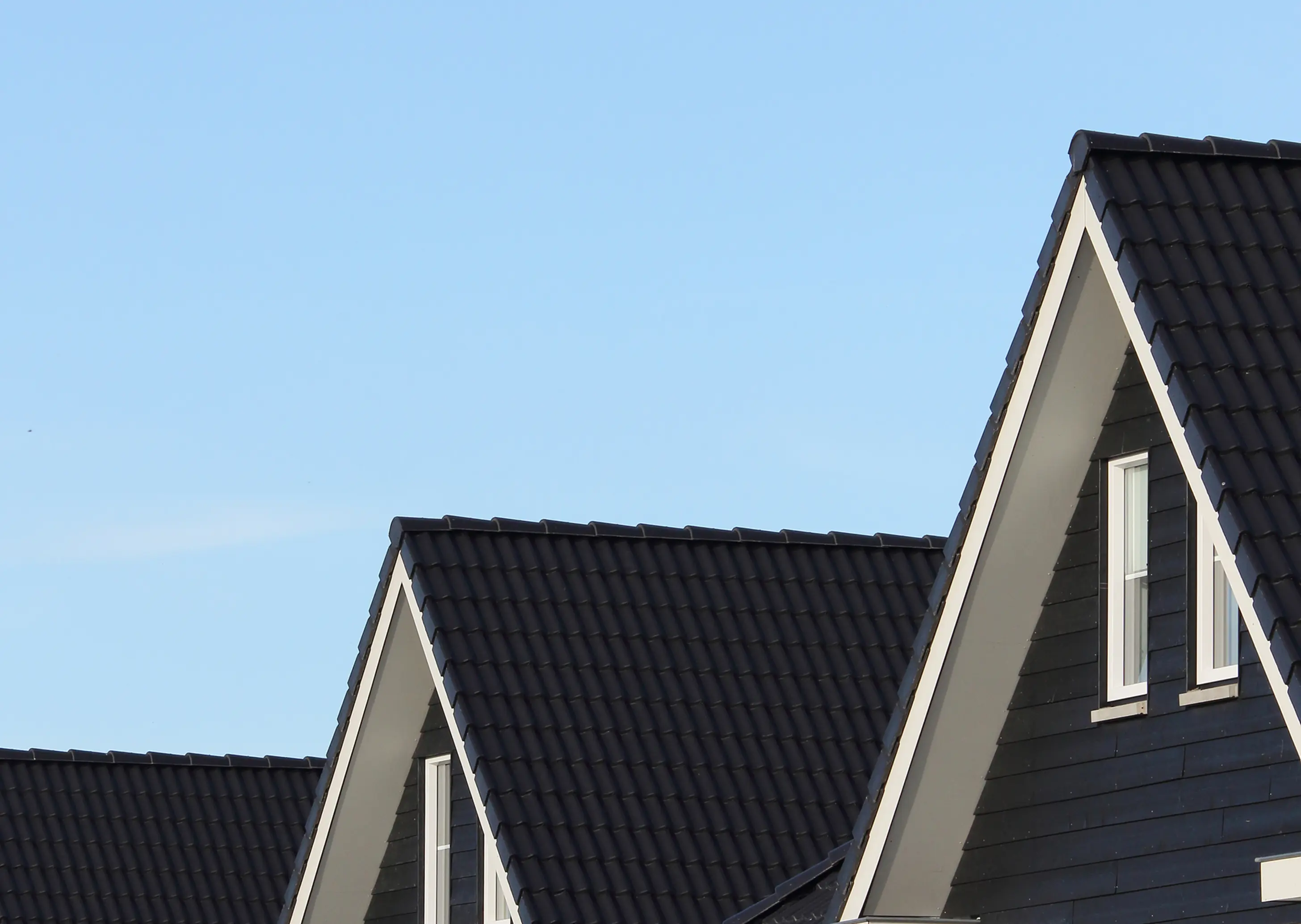 Close-up of three house rooftops with black tiles and white-framed windows under a clear blue sky.