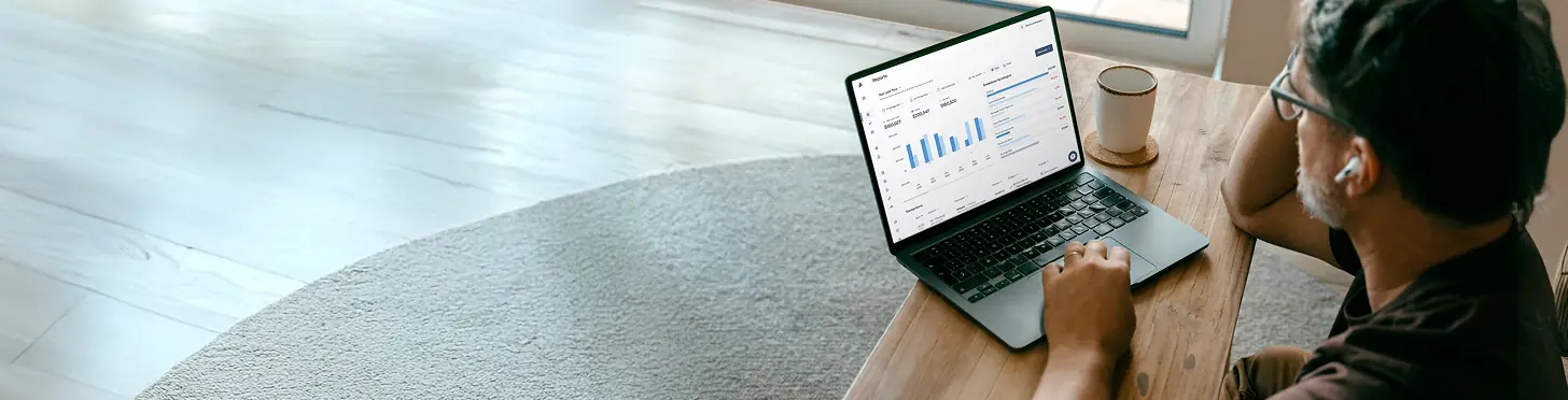 Man wearing glasses and earbuds working on a laptop with finance or sales reports displayed, sitting at a wooden table near a window.
