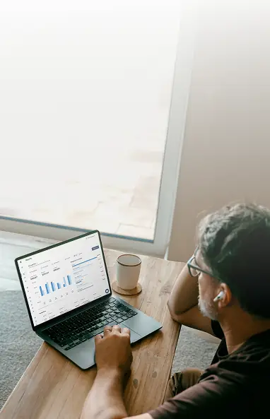Man wearing glasses and earbuds working on a laptop showing graphs and data at a wooden table near a window with a cup of coffee.