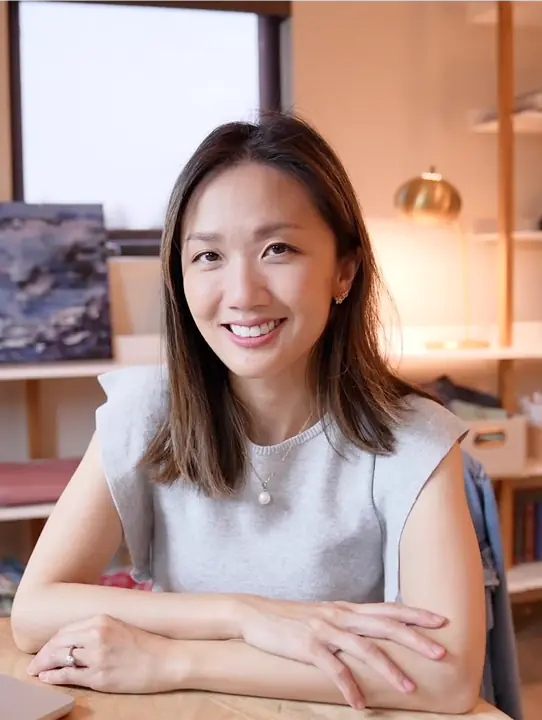 Smiling woman with shoulder-length hair sitting at a desk with arms crossed, wearing a gray sleeveless top and pearl necklace in a warmly lit room.