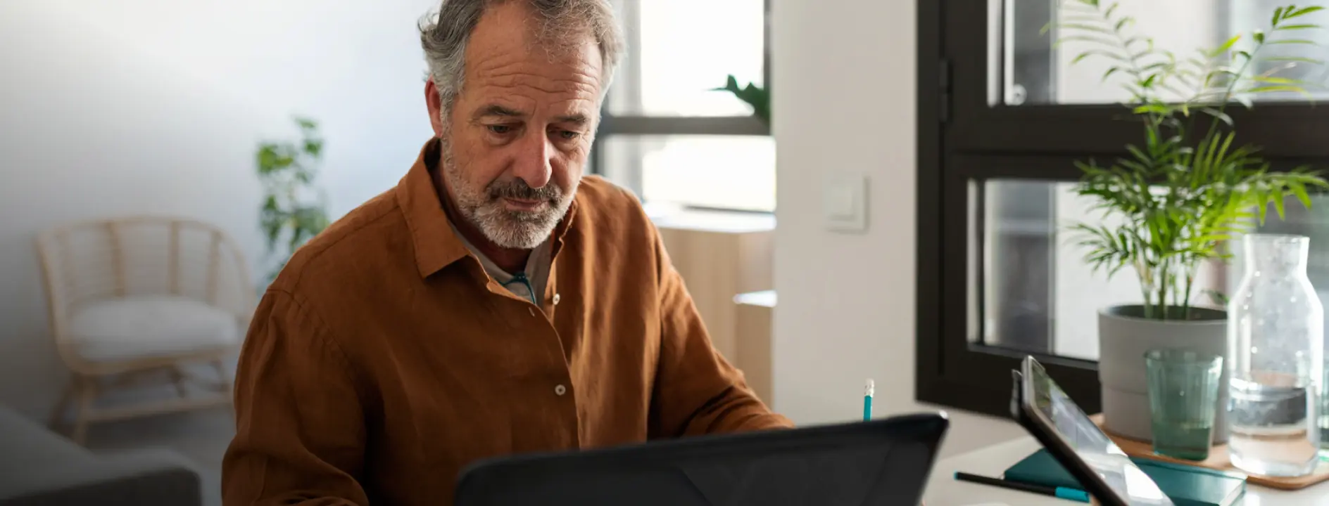 Middle-aged man in a brown shirt focused on his laptop at a desk with plants and a glass water bottle nearby.