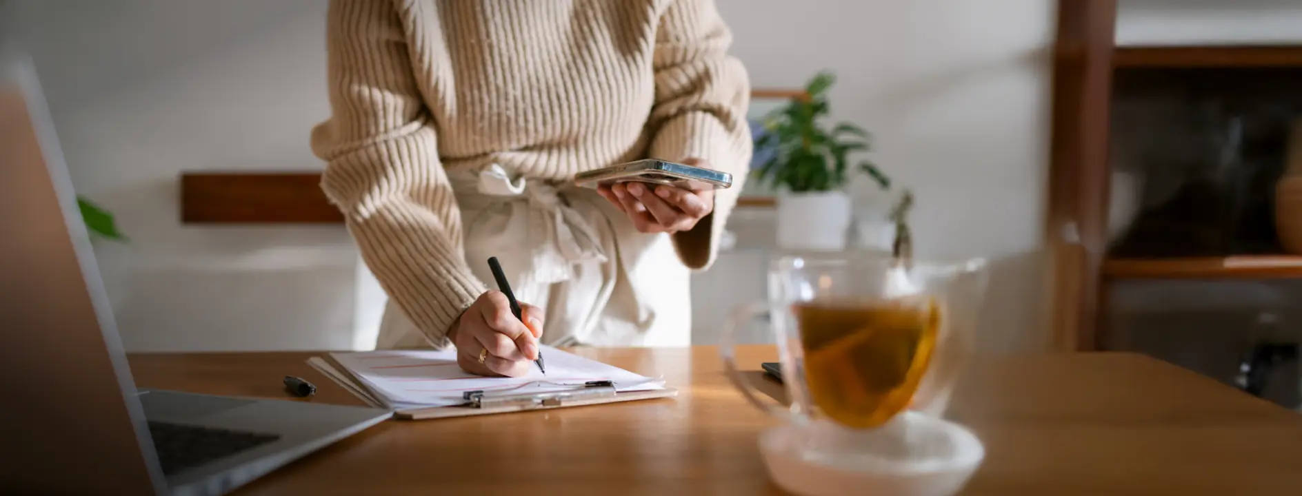 Person wearing a beige sweater writing on a clipboard while holding a smartphone, with a laptop and a glass cup of tea on the wooden table.