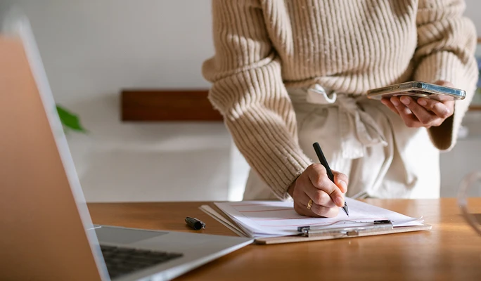 Person in beige sweater and apron writing on documents with a pen while holding a smartphone, near a laptop on a wooden table.