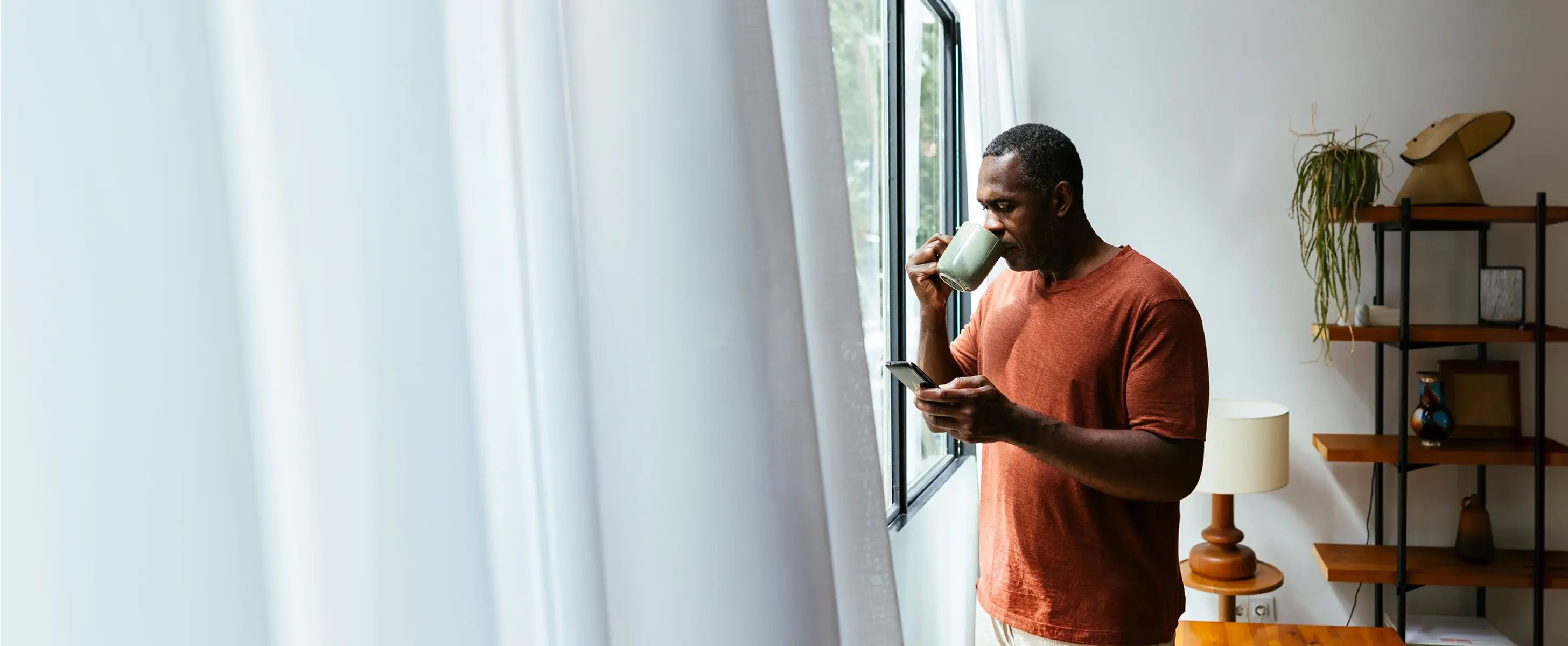 Man in an orange shirt drinking from a green mug while looking at his phone by a window in a bright room.