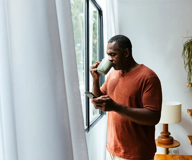 Man in an orange shirt stands by a window, drinking from a light green mug and holding a smartphone.