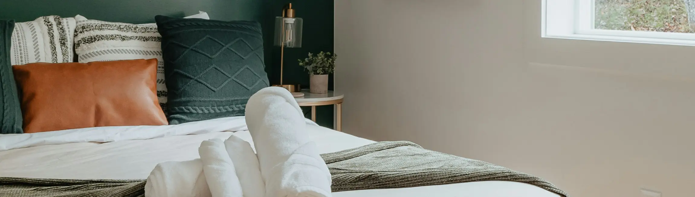Cozy bed with white sheets, textured pillows, rolled white towels, and a small bedside table with a lamp and plant near a window.