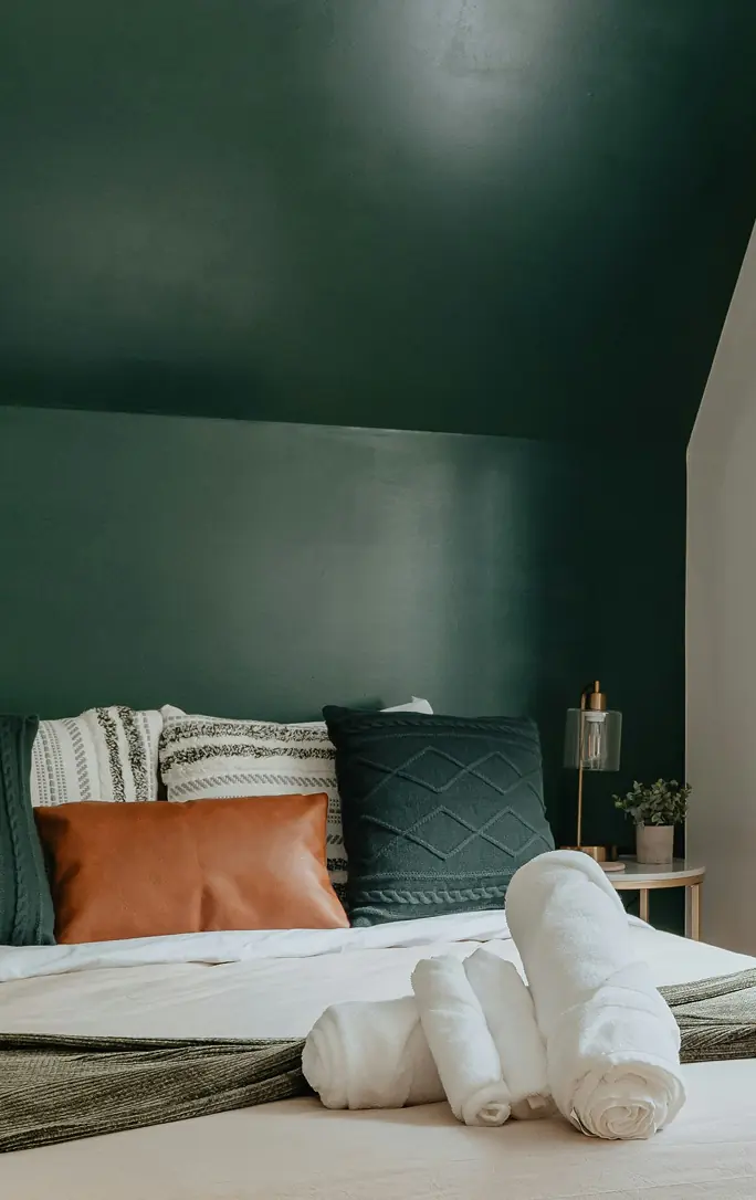 Bed with white linens and assorted pillows against a dark green wall, with rolled white towels in the foreground and a bedside table holding a lamp and small plant.