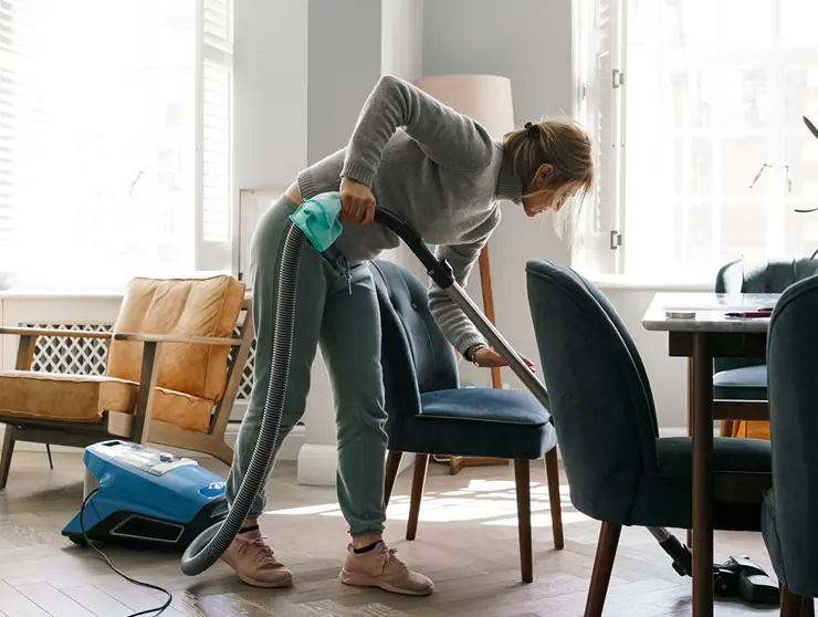 Woman vacuuming under a dining chair in a bright, modern living room with wooden floors and natural light.