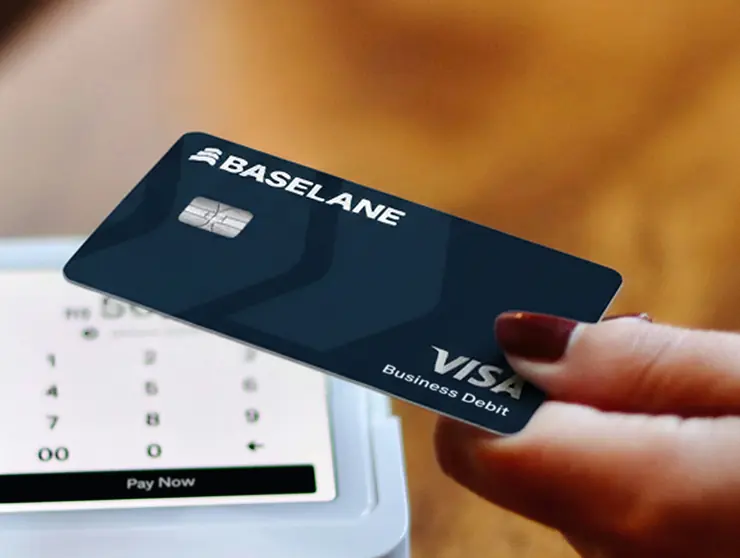 Hand with dark red nail polish holding a Baselane Visa Business Debit card near a payment terminal screen showing a keypad and a Pay Now button.