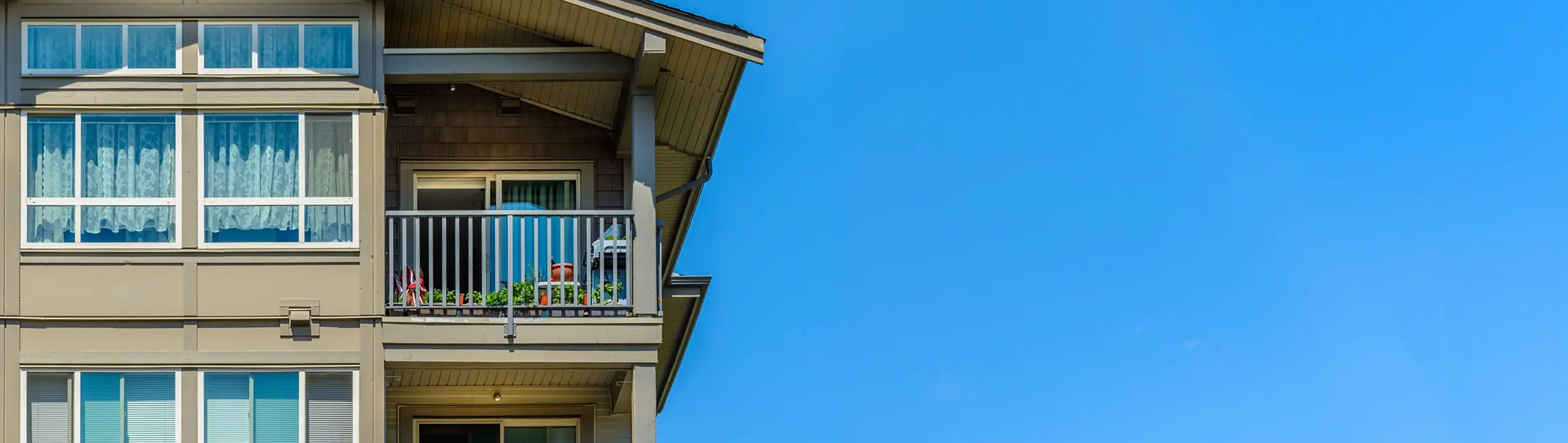 Corner section of a beige apartment building with a balcony and windows, under a clear blue sky.