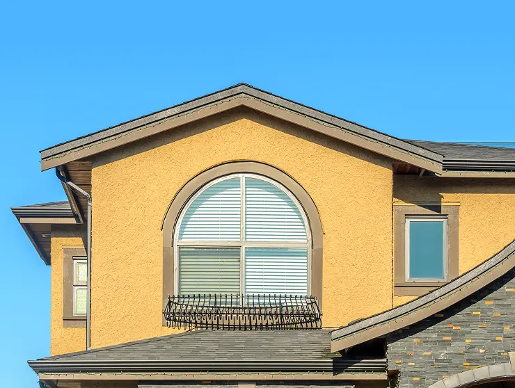 Yellow stucco house exterior with a large arched window and a small rectangular window under a clear blue sky.