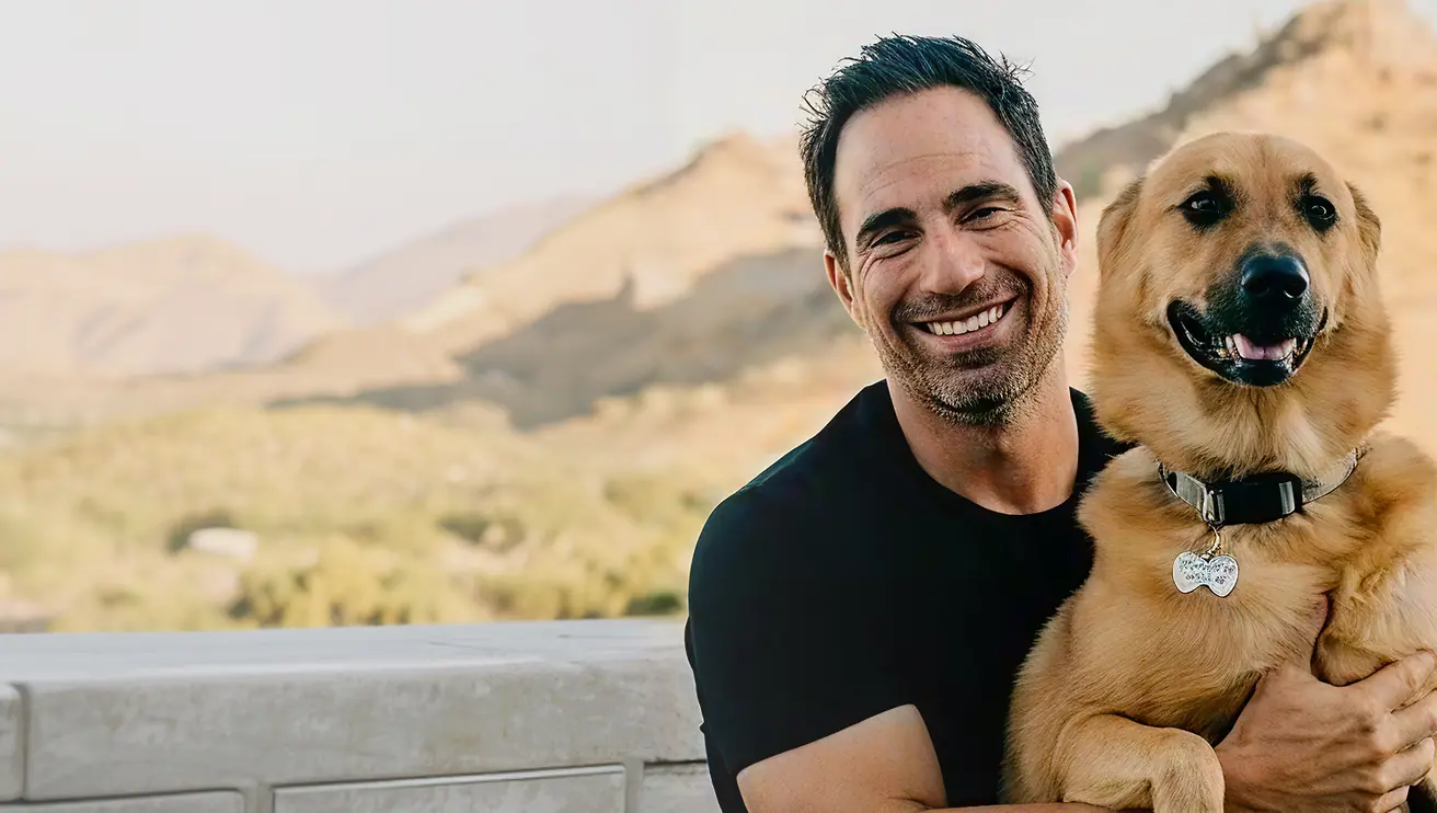 Smiling man in a black shirt hugging a happy golden retriever dog outdoors with mountains in the background.