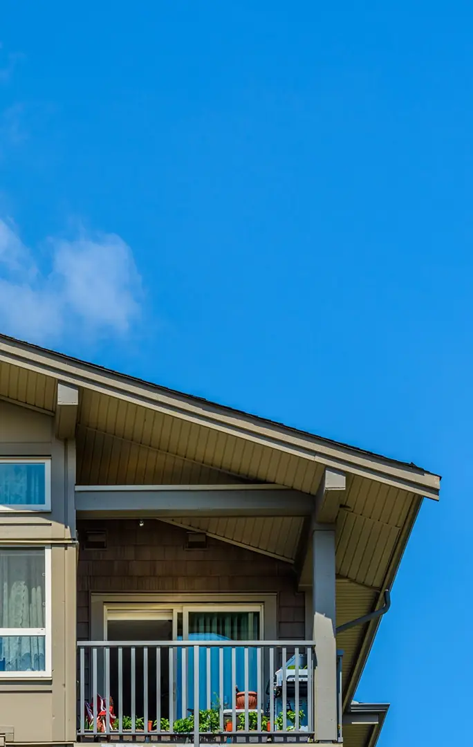 Upper corner of a beige house with a small balcony, sliding glass door, and clear blue sky background.