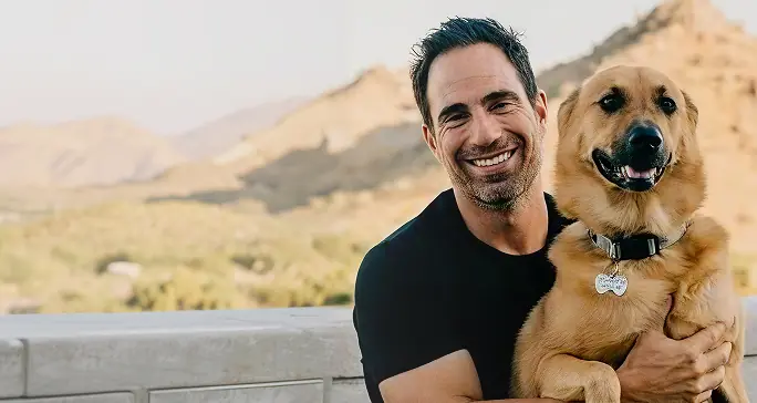 Smiling man in a black shirt holding a happy golden-brown dog outdoors with mountains in the background.