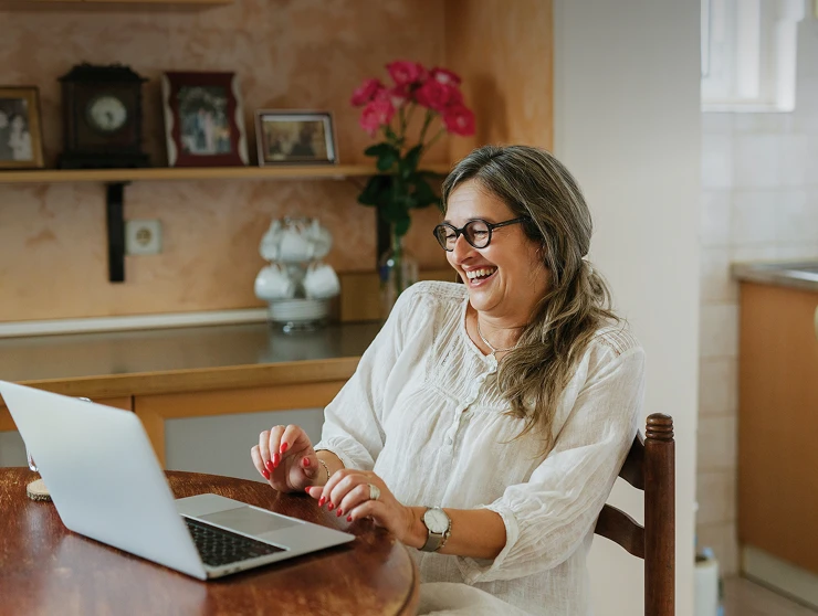 Smiling woman with glasses and long hair working on a laptop at a round wooden table in a cozy kitchen.