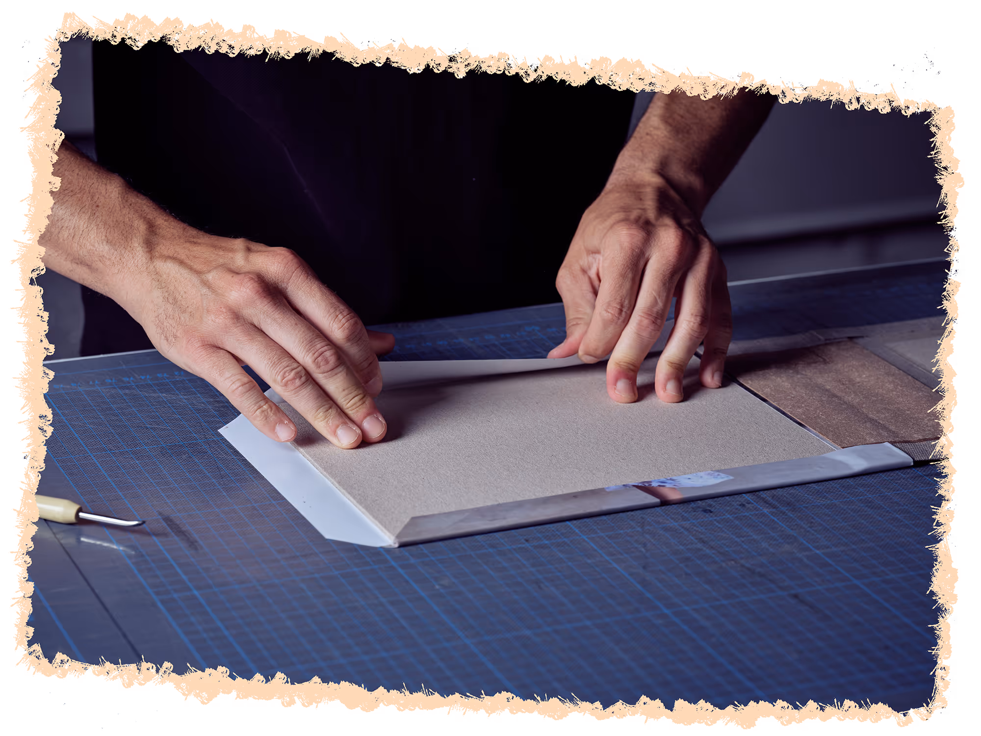 Close-up of hands folding a piece of cardboard on a cutting mat with a craft tool nearby.