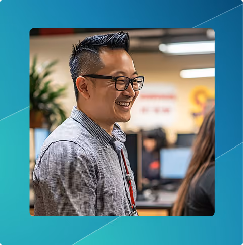 Smiling man with glasses and a checkered shirt in an office setting with computer monitors in the background.