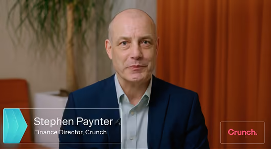 A middle-aged bald man in a dark blue suit and light shirt looks at the camera, identified as Stephen Paynter, Finance Director at Crunch, with a green and white name tag and a red Crunch logo on the right side.