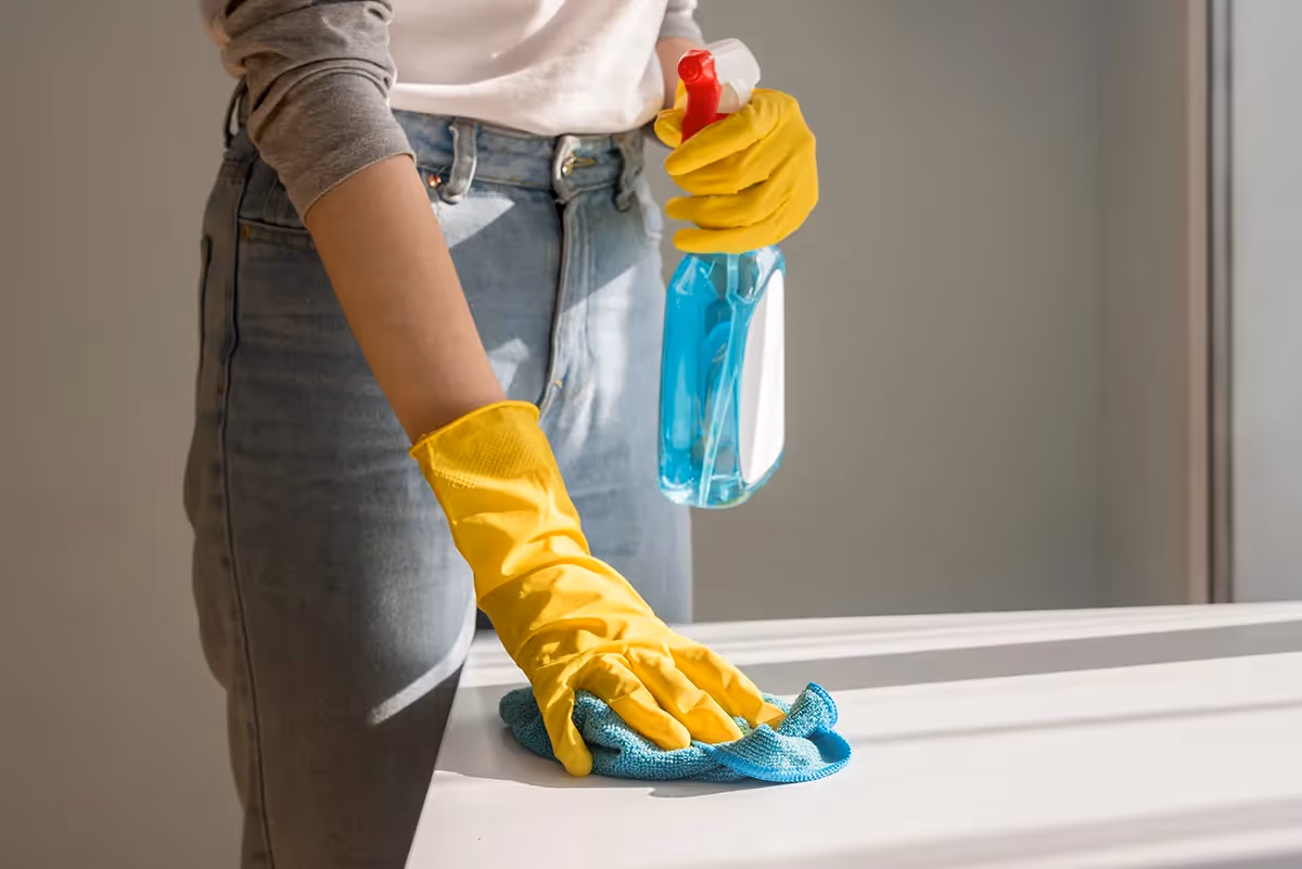 Woman wearing yellow gloves cleaning surface with spray bottle and cloth in sunlight, Regular Cleaning Services in London