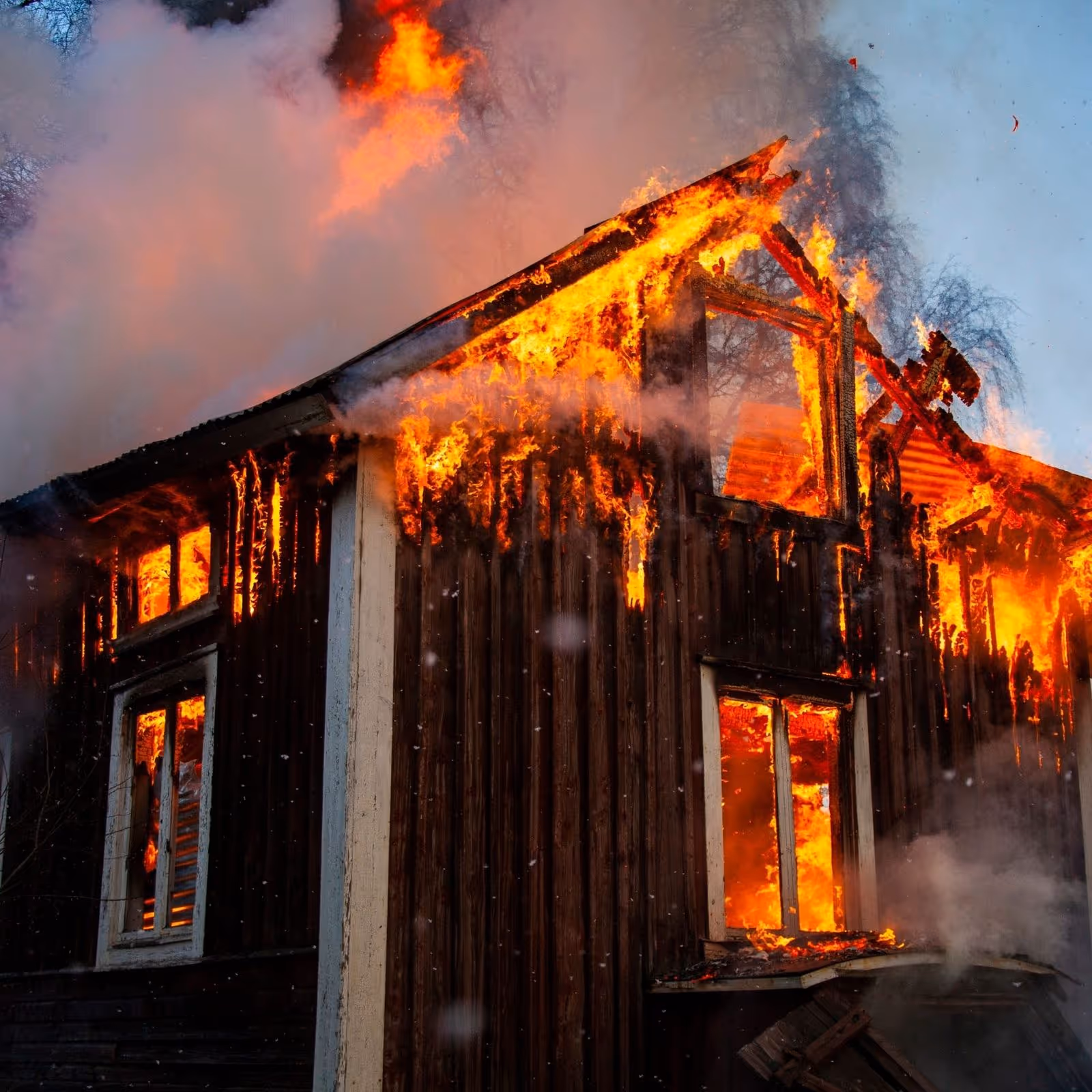 Wooden house engulfed in intense orange flames with thick smoke at dusk or dawn.
