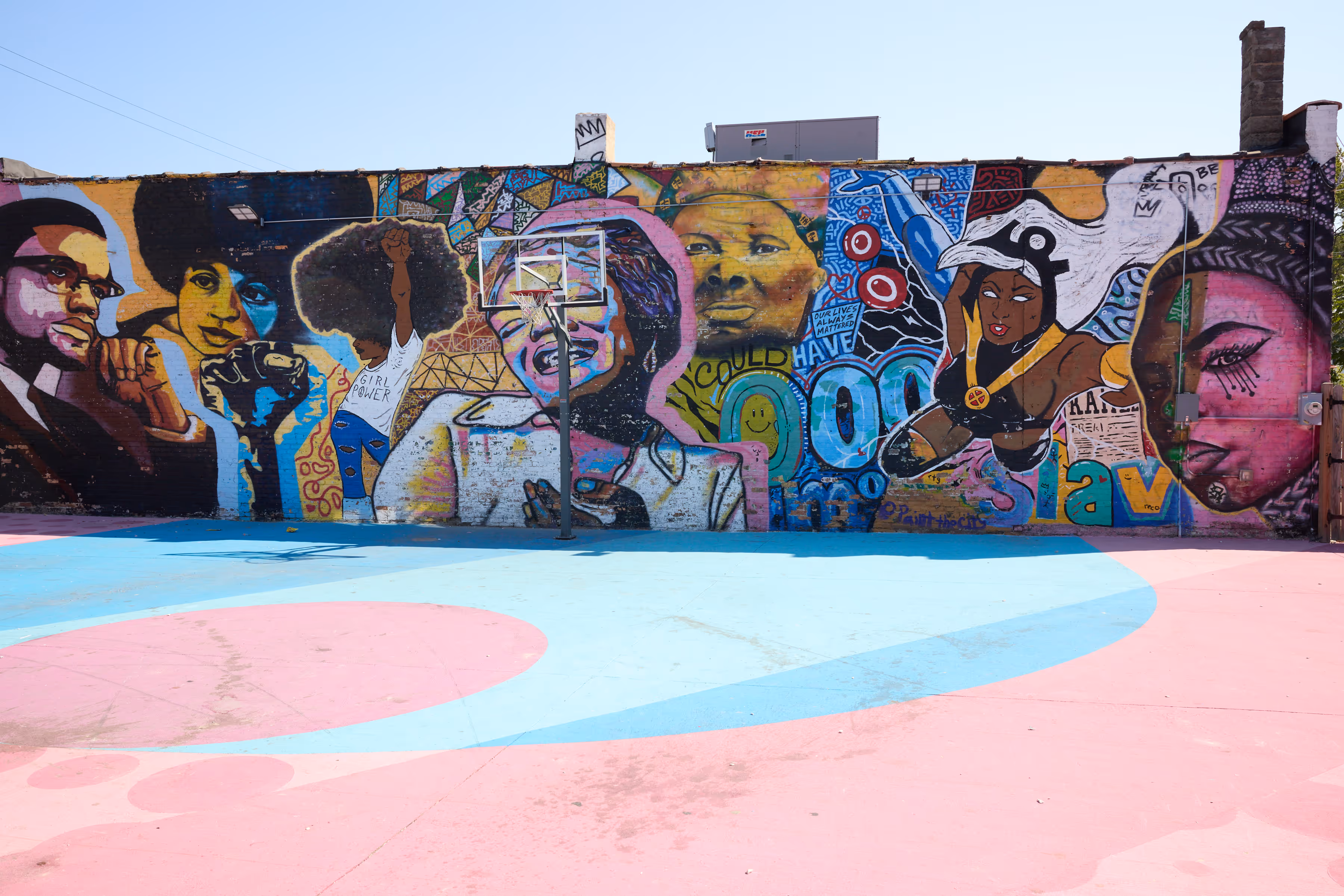 Colorful mural on a basketball court wall featuring portraits of Black activists, a raised fist, a woman with 'Girl Power' shirt, and comic-style superhero.