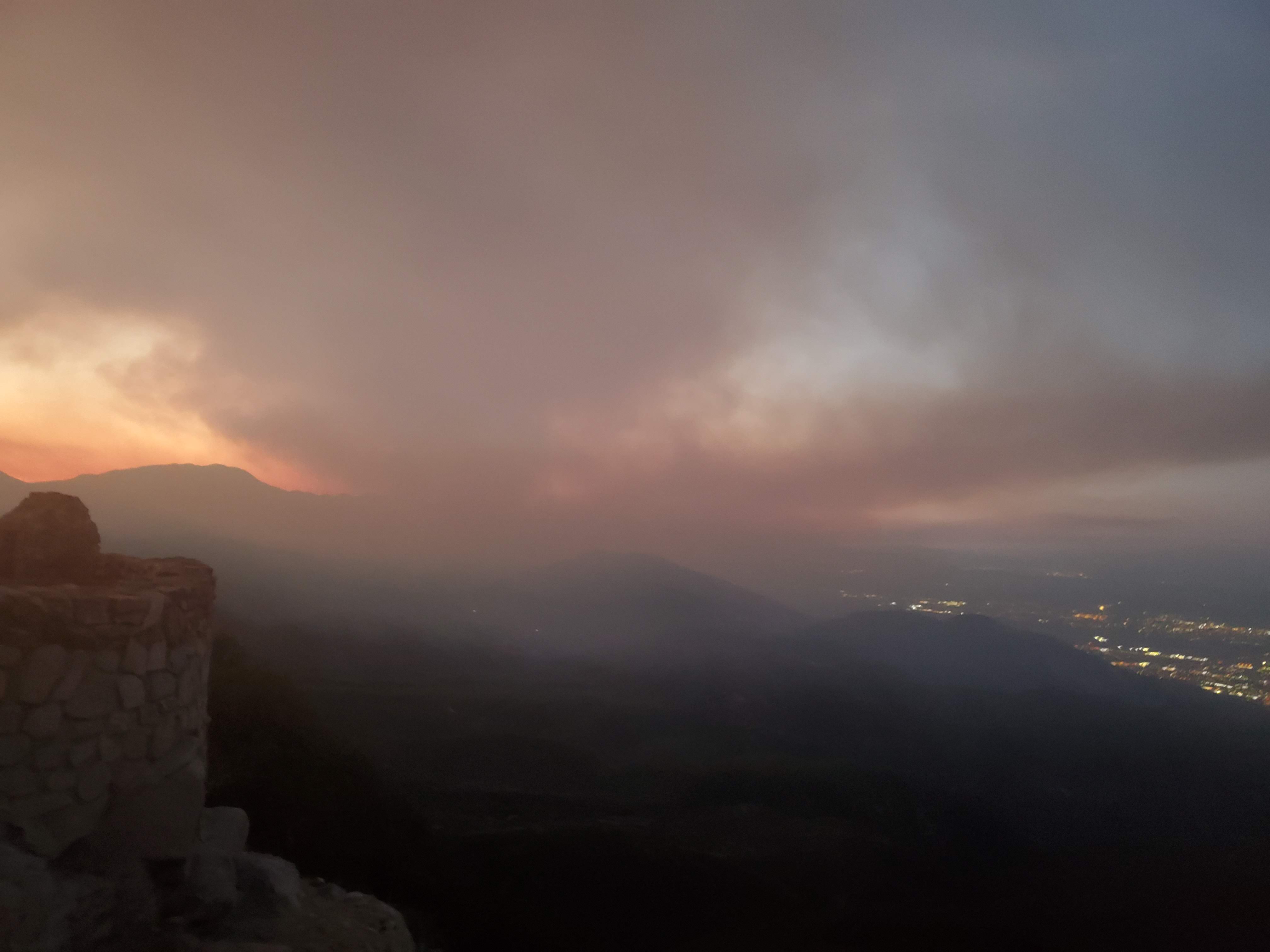 Extreme dark smoke clouds the sky over a scenic mountain view with a wildfire in the distance