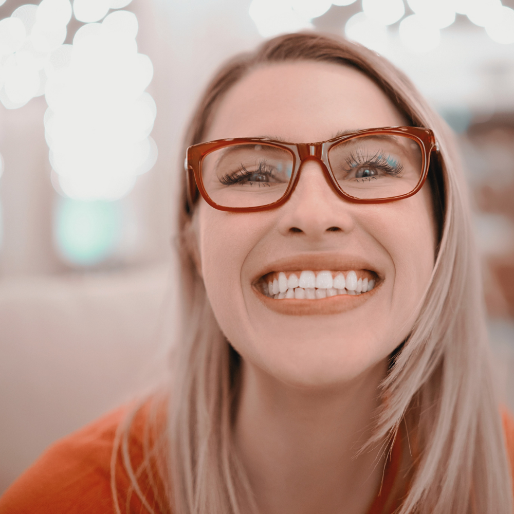 Woman with red glasses and blonde hair smiling and looking at the camera, showing off teeth