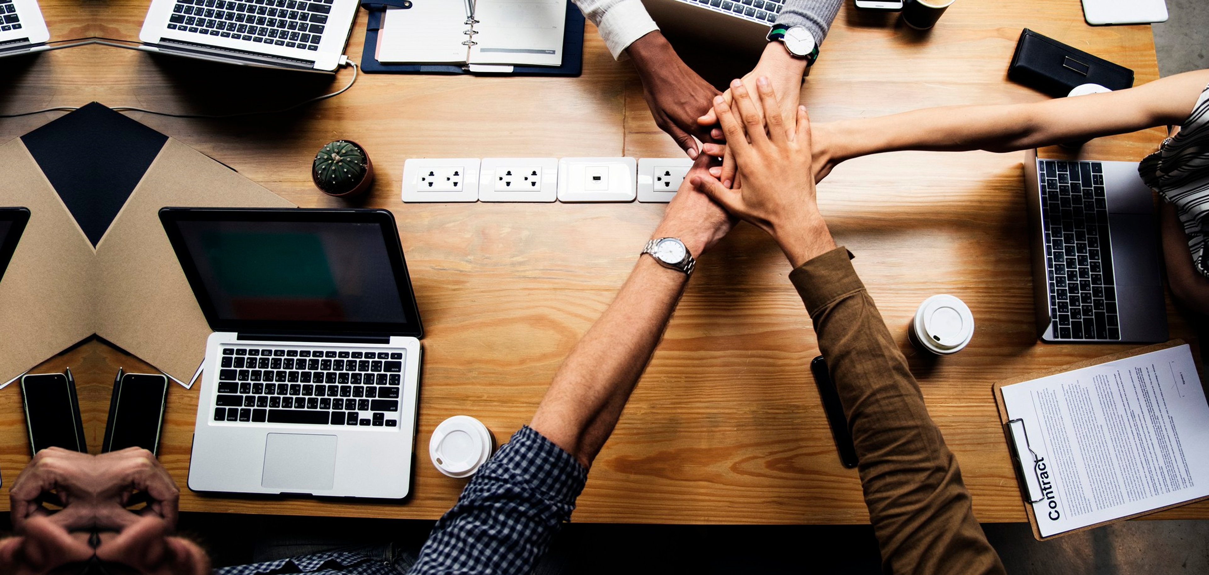 Top view of diverse team stacking hands over a wooden table with laptops, phones, coffee, and contract.