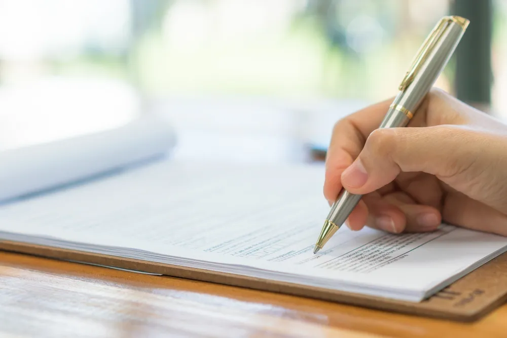 Hand holding pen poised to write on a document with printed text on a wooden table.
