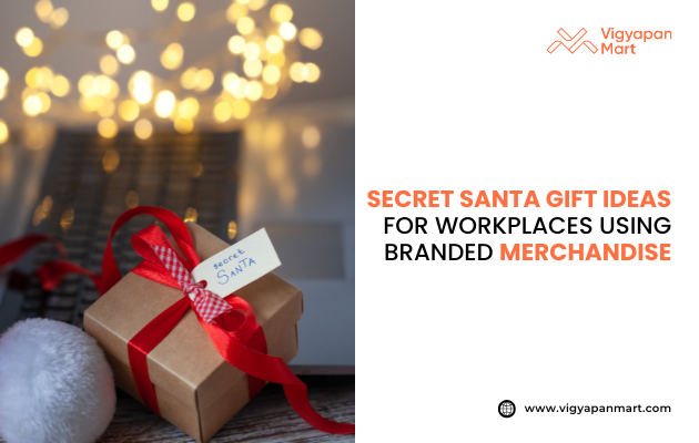 Small gift box with red ribbon and "Secret Santa" tag placed on a desk with holiday lights in background. Text reads "Secret Santa Gift Ideas for Workplaces Using Branded Merchandise." Vigyapan Mart logo top right, website bottom right. Focus on workplace holiday gifting.