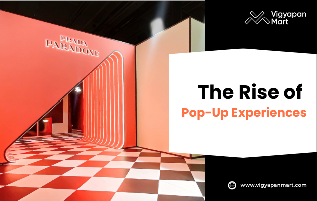 Modern pop-up store with bold red and white design featuring a geometric checkered floor, illuminated tunnel entrance, and signage reading “Prada Paradoxe.” The right side of the image includes a promotional banner for Vigyapan Mart with the headline “The Rise of Pop-Up Experiences” and the website URL www.vigyapanmart.com