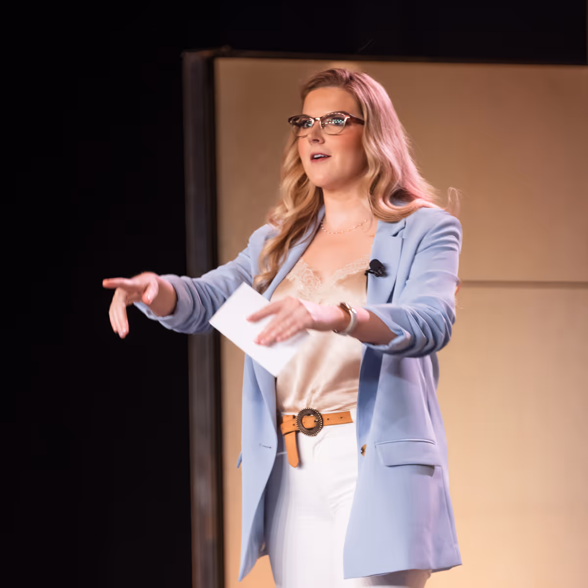 A woman in a light blue blazer and glasses speaking on stage with outstretched hands, holding a note card.