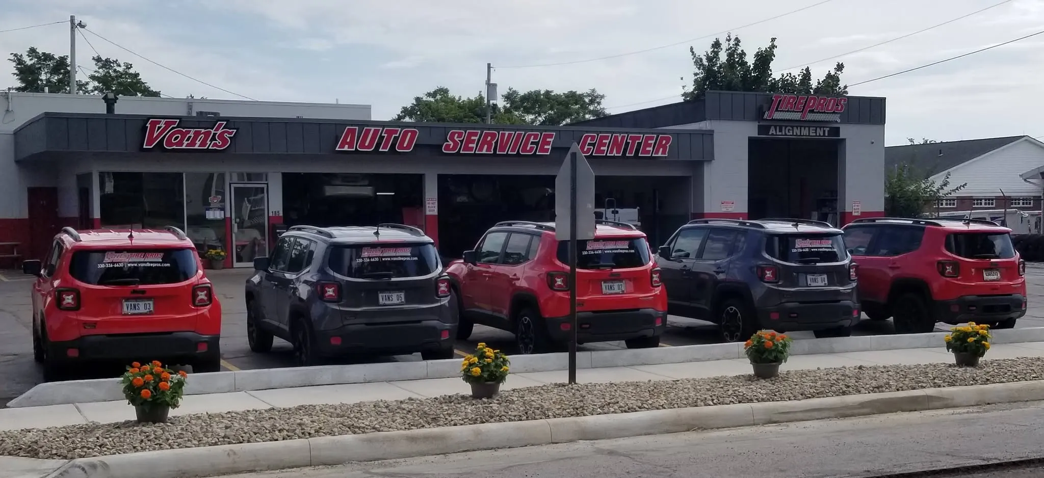 A row of vehicles parked outside Van's Auto Service Center, with vibrant flowers in planters and a clear blue sky above.