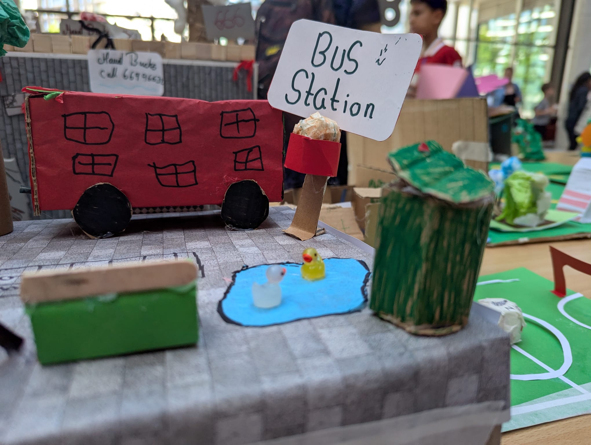 A close-up photo of a model bus station. A paper duck pond and cardboard
recycling bin are next to the bus-stop bench.