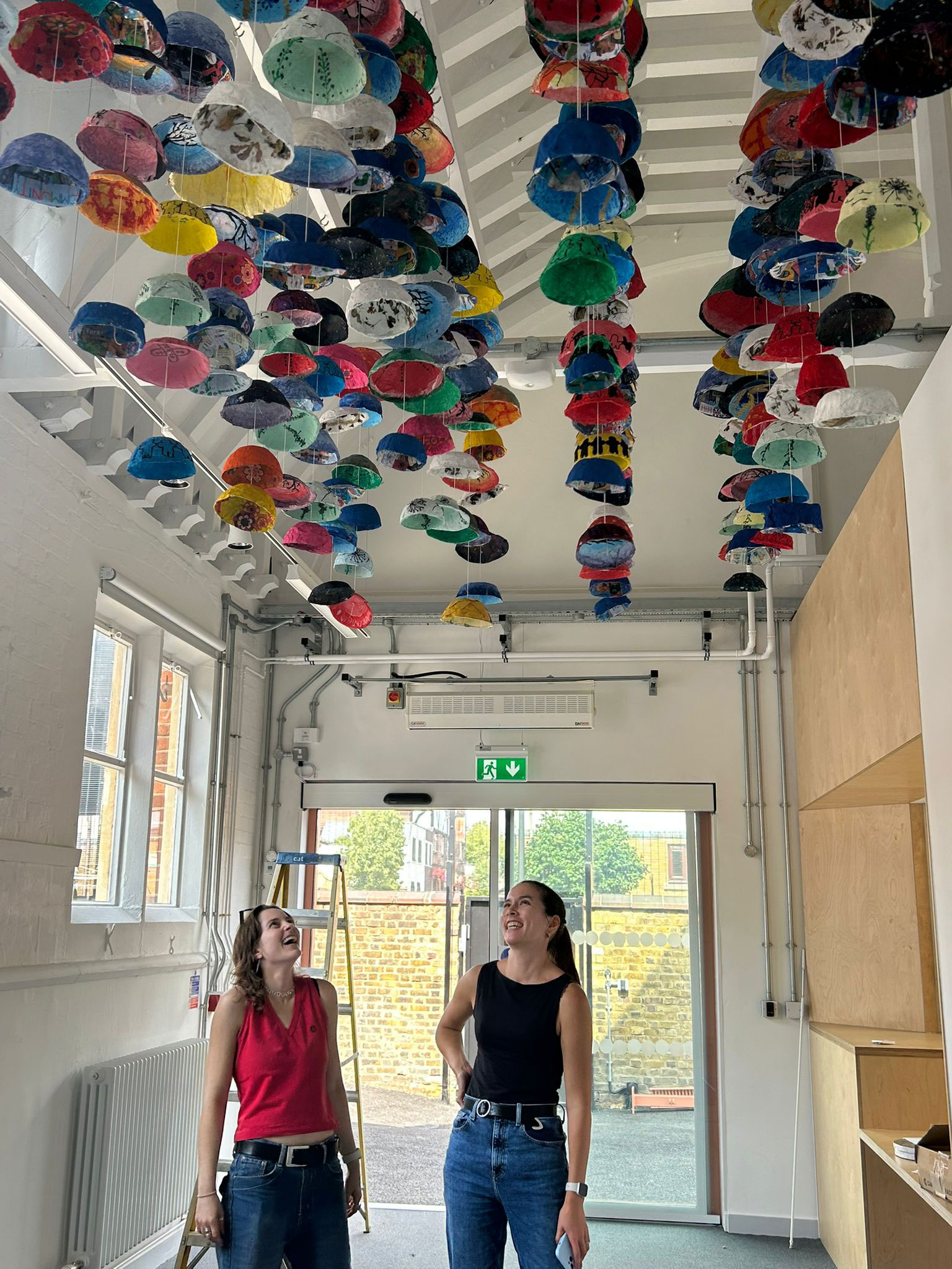 Bessie and Sophie pose below the completed installation. The installation
takes the form of many bowls hanging from the ceiling creating an ‘umbrella’ effect.
The installation in the entrance to one of the buildings at the school.