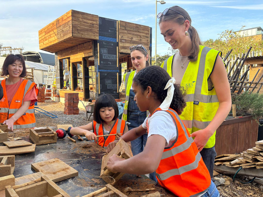 Ella assisting children in handmade brickmaking. They wear hi-vis while
standing in front of what will be the office at the Triangle Site, currently clad in wood
and handmade brick.
