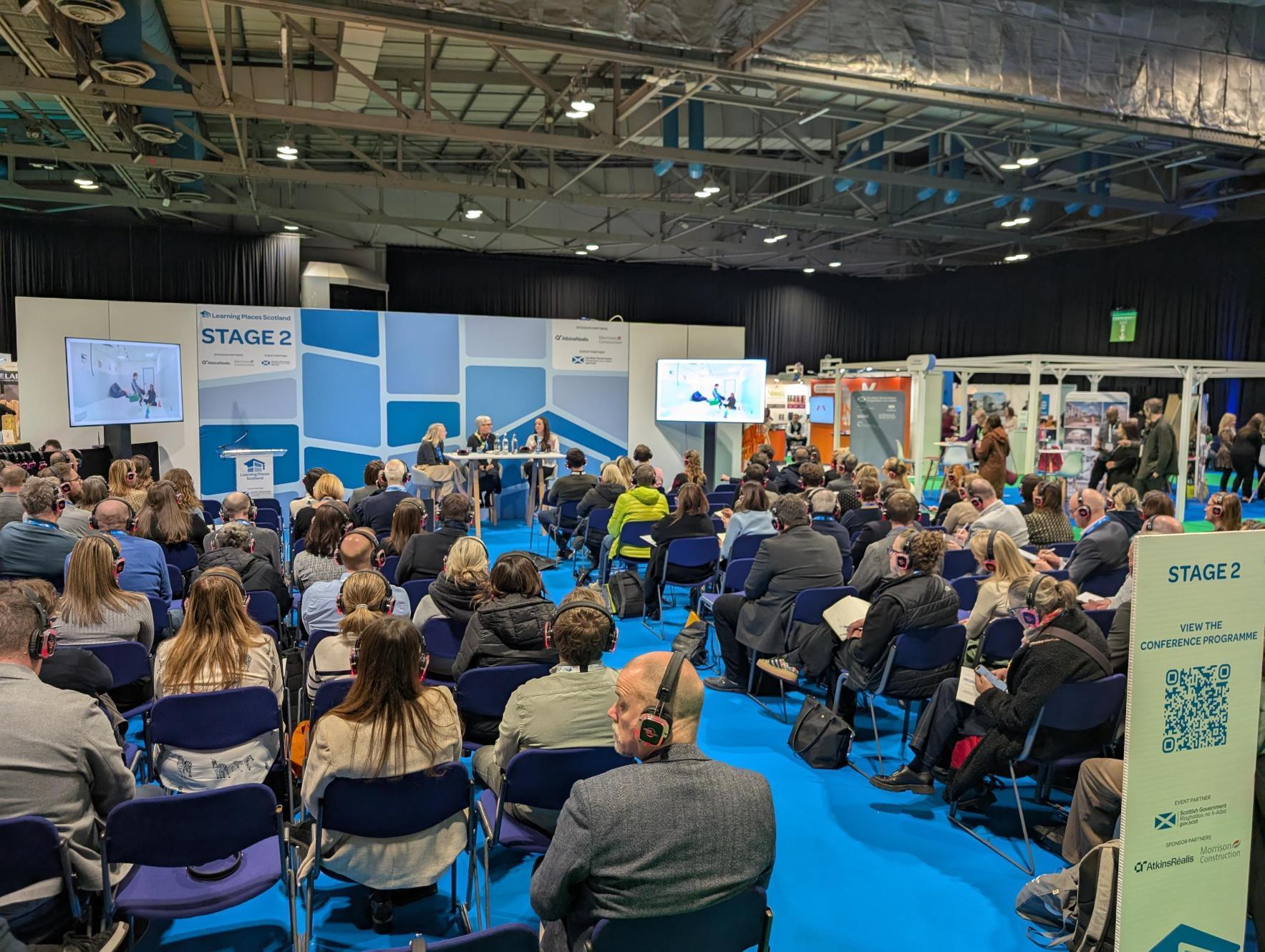 Left to Right: Claire Barton, Claire Jackson and Jennifer Freeman, they are sat
at a table on the stage at education estates. Behind them is a background of various
shapes in hues of blue and grey. Above them, text reads ‘Inclusive Estate at Learning
Places Scotland’