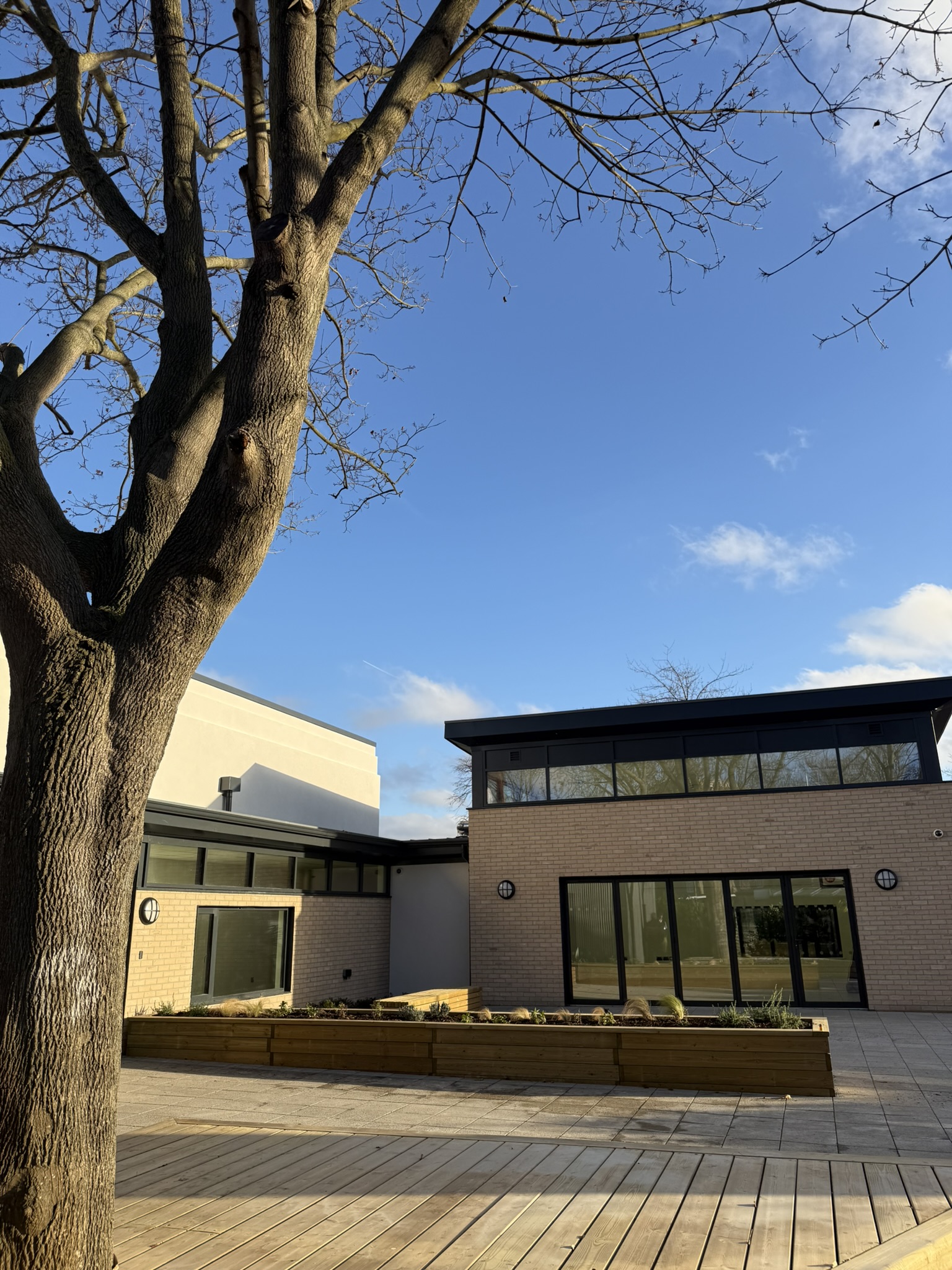A photo of the recently completed Rectory from the
outside. The building has a brick facade with a black-framed
ceerestory window below the roof. To the left is a tree and
there is a strip of planters filled with young plants infront of the
building.