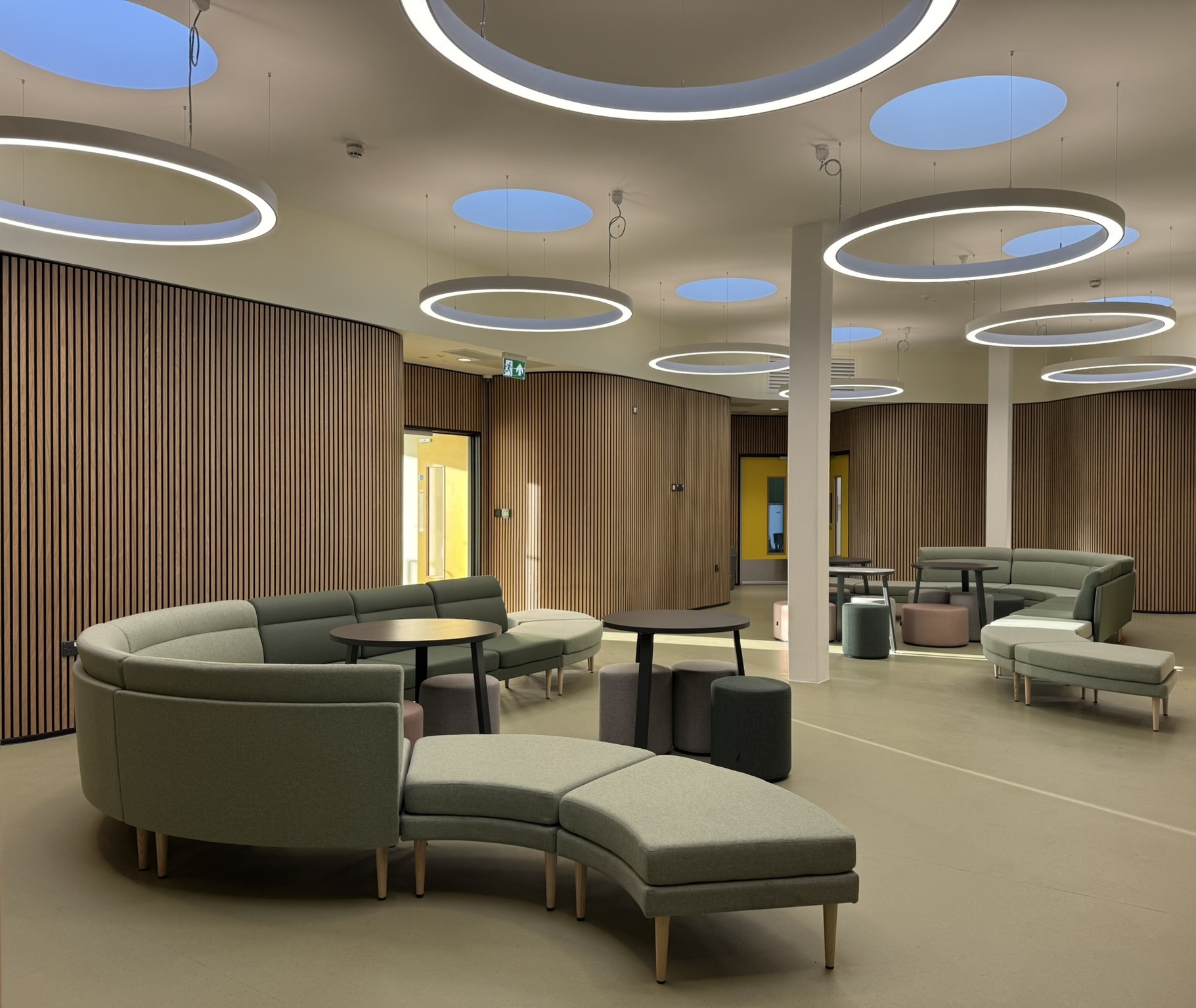 The interior common space inside the building. The
walls are covered in a light-wood panelling. The ceiling has
scattered circular lights framing circular skylights allowing for
natural light. In the room there are modular-light green sofas
alllowing for a flexible layout.