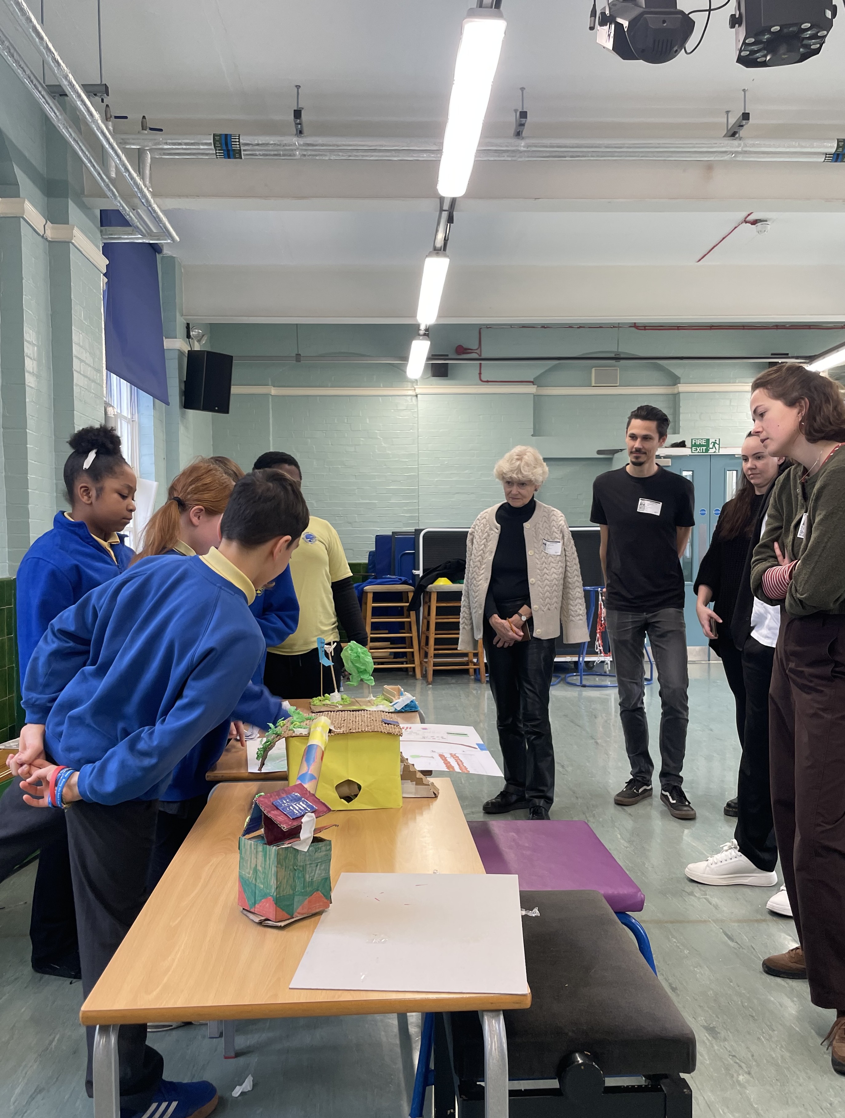 Students to the left present their work to designers, who stand on the right. Between them is a table with the students models on it.