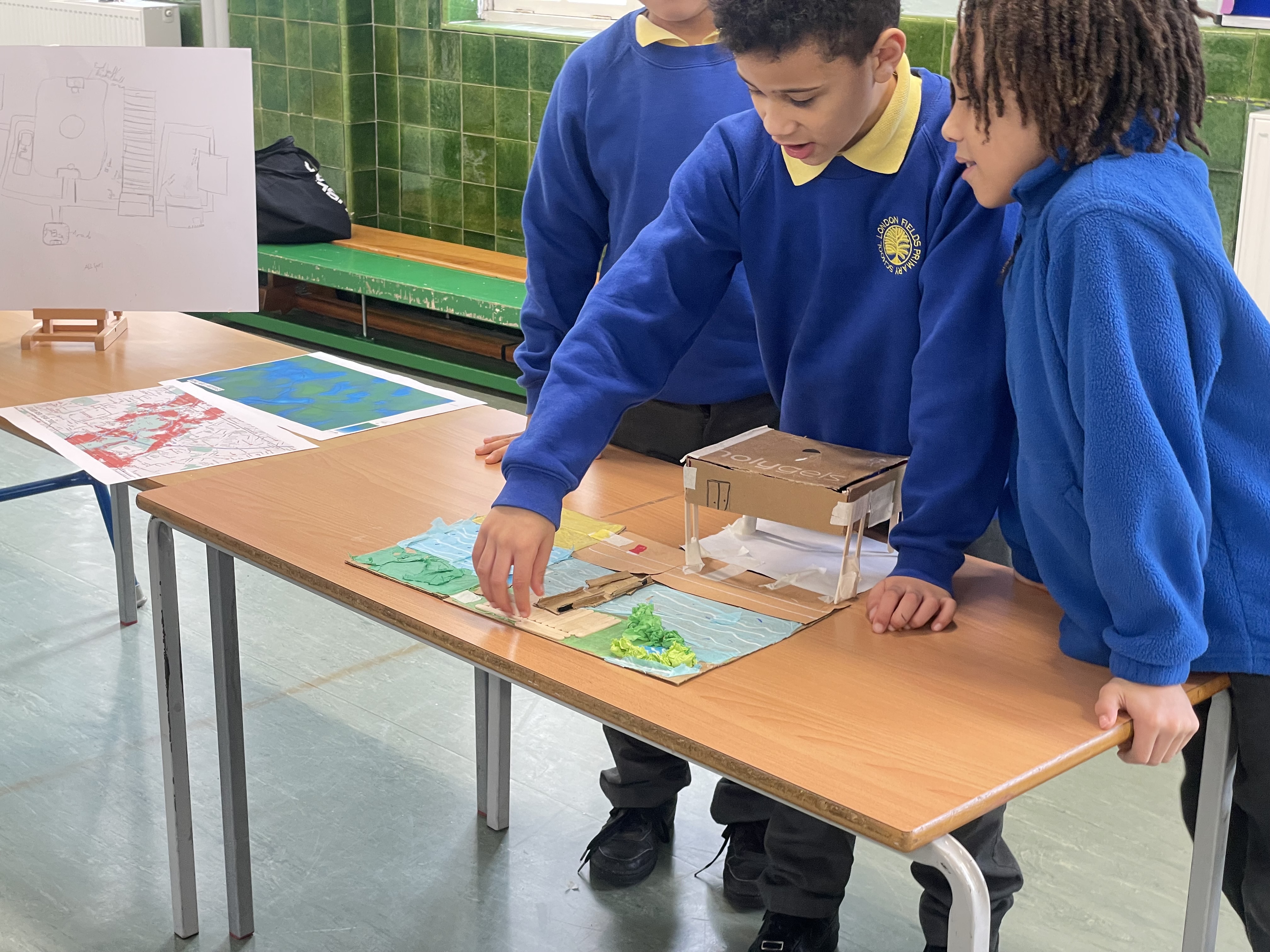  Three students share their work, stood behind the table where they present.