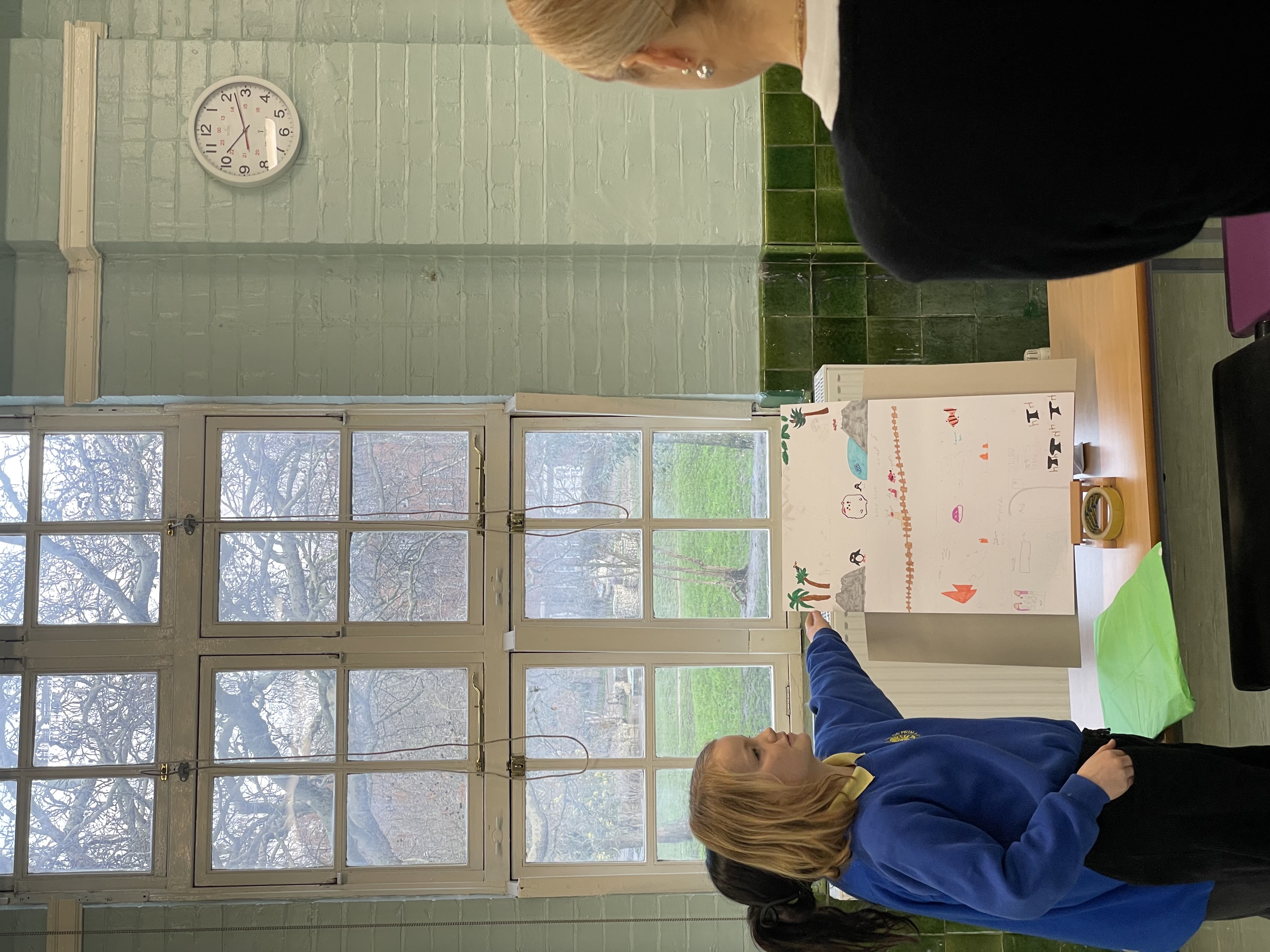 Two students point toward their model while presenting their work. The table display is in front of a window.