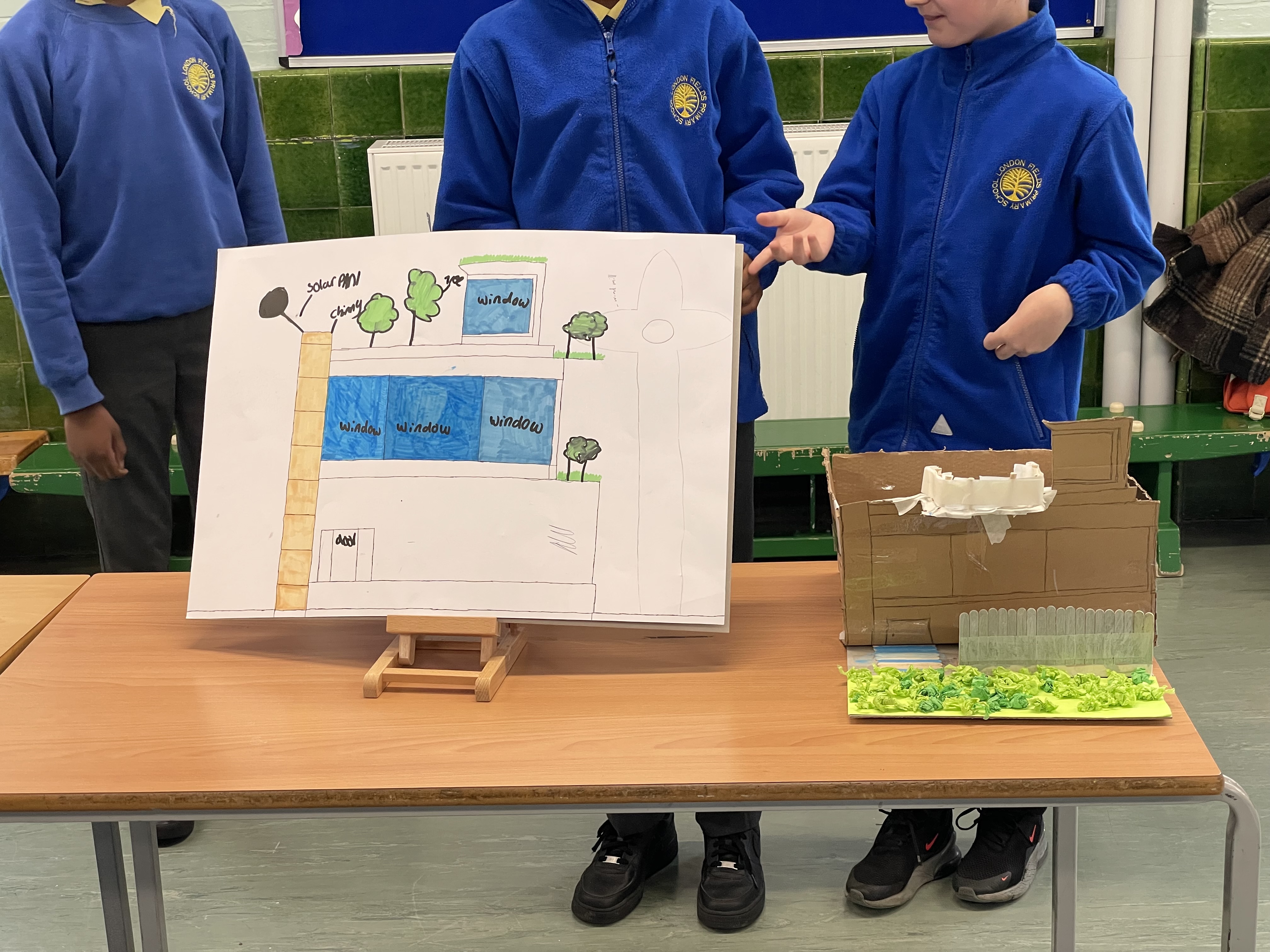 The students plan drawing next to their cardboard model. The model includes vegetation and a two storey building.