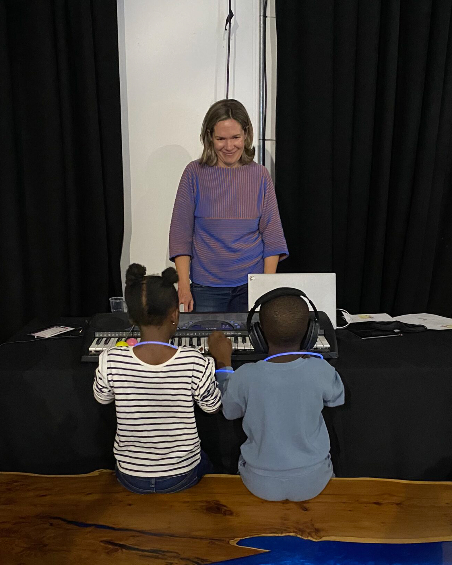  Emma stood behind a keyboard facing forward and the backs to two small children playing the keyboard of whom we can only see their backs. They are sat on a stool. There is no text on this image.