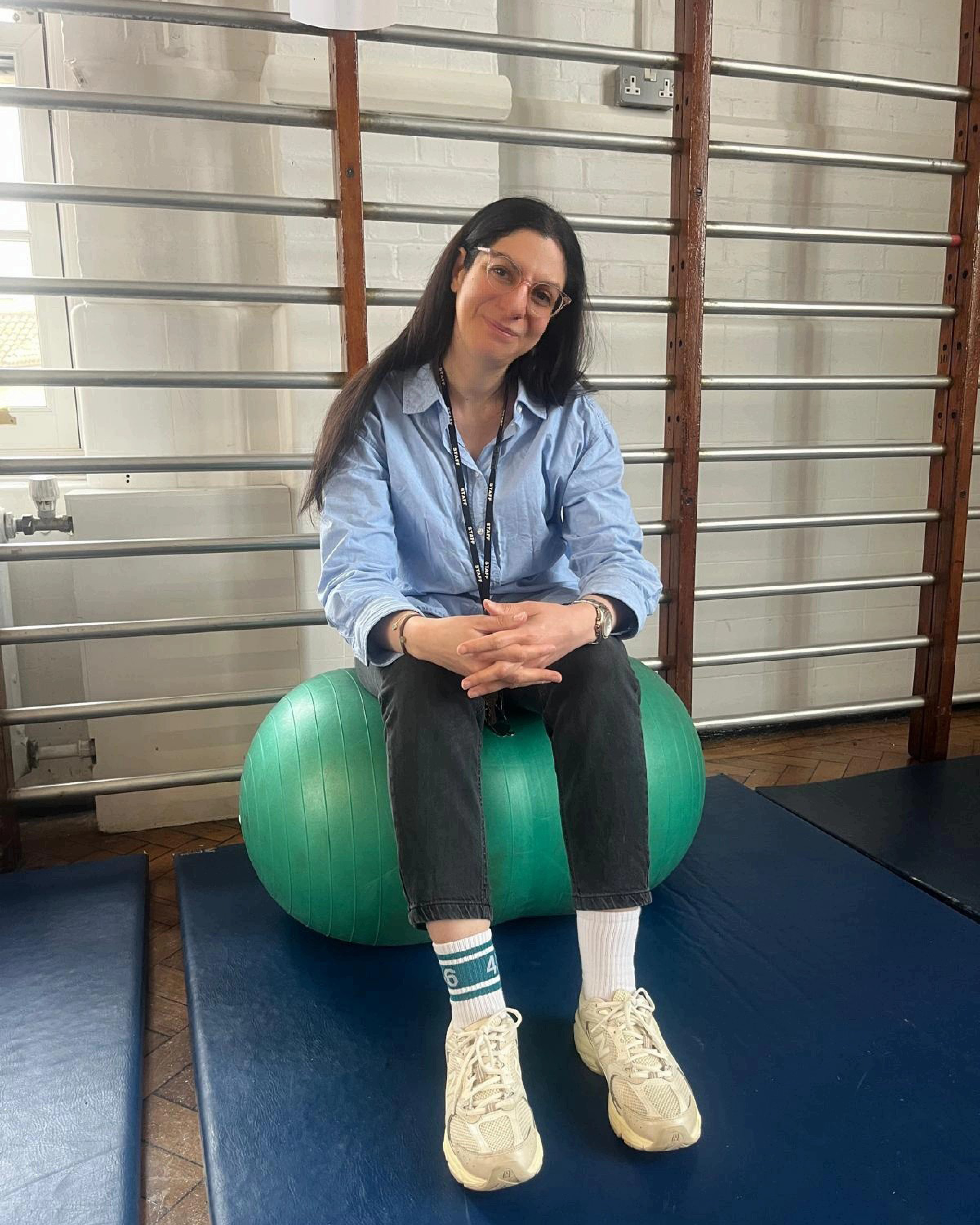 An image of Heba in the school hall, sitting on a green yoga ball. She wears black trousers and a blue button up top.