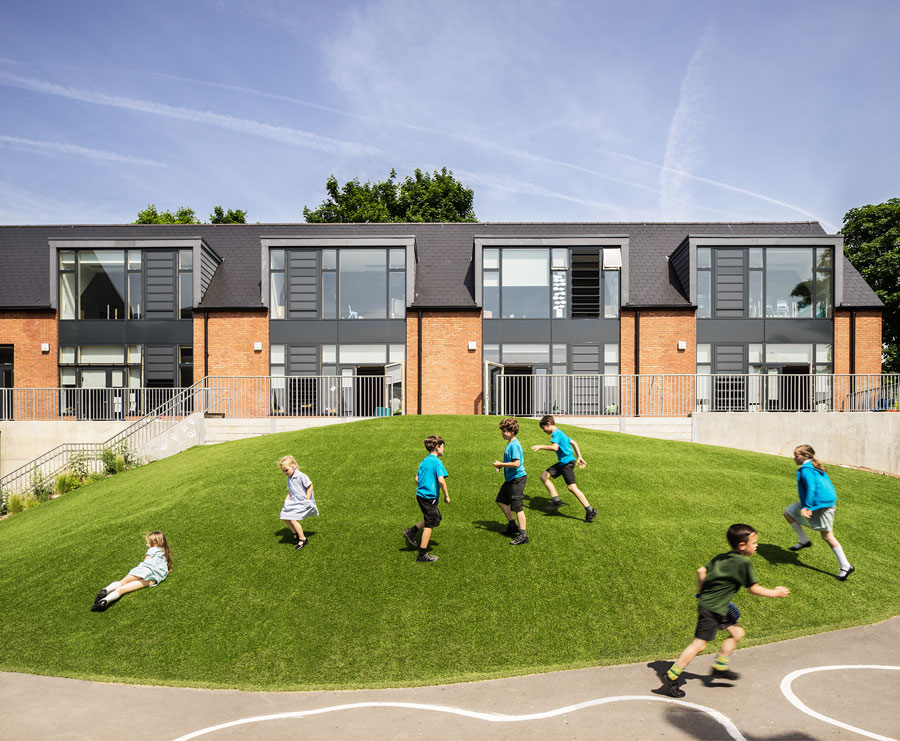 The new lower school accommodation. The building takes the form of a terrace, with large outdoor space outside each classroom. In the foreground students play on a grassed mound.