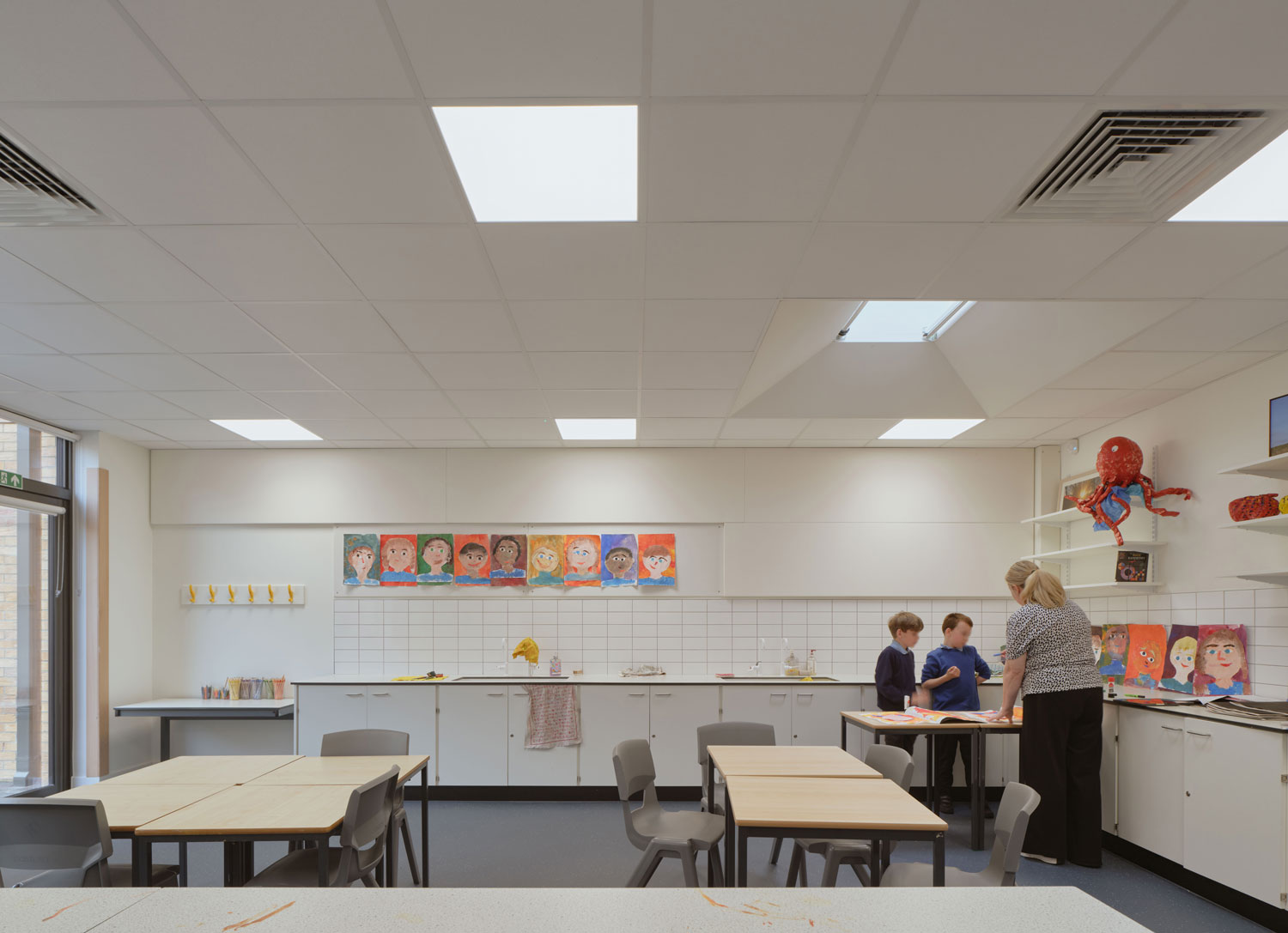An art classroom at Hawthorns, paintings line the wall with two students and a teacher to the right