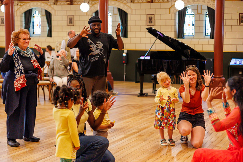 A cultural dance workshop in Connaught Hall. To the left is a woman wearing a Sari, teaching moves to a group of children and adults.
